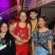 Four people pose together in a public venue, smiling and relaxed as they gather for a photo. In the center, 9a stands out in a red tank top featuring illustrated wolves, with long red-and-black hair and a confident, they wear glasses and are smiling. To their left, a person wears a floral off-shoulder top and smiles warmly, behind them a man in a white t-shirt with short dark hair is smiling to the camera. Another individual holds a rolled-up poster and a magazine, they are smiling while wearing a black shirt with a colorful print, they have long black hair and wear glasses and a necklace.