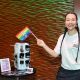 A person in long black box braids is waving a small pride flag in their right hand, they’re smiling, wearing pink earrings, red lipstick, a white T-shirt and black bottoms. On their side there’s a small setup of earrings and stickers being displayed, as well as a small frame with payment methods.