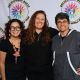 Three sign language interpreters stand together in front of a white backdrop featuring circular logos that read “DECOLONIZED BEATZ INDIGENOUS WORLD PRIDE”. All three are smiling warmly and dressed casually. The interpreter on the left wears a black outfit with colorful accessories and has their arm around the person in the middle, who is dressed in a black top and leans in with a cheerful expression. On the right, the third interpreter wears a gray jacket with white trim and smiles with relaxed posture.