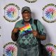 A man stands smiling in front of a white backdrop featuring the repeated circular logo and text “DECOLONIZED BEATZ INDIGENOUS WORLD PRIDE.” He wears a green patterned polo shirt, jeans, a green cap, and large tinted glasses. A black shoulder hangs from his body, and he holds a small rainbow flag with feathers in the center.