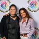 Two women stand side by side in front of a white backdrop featuring the circular logo and text “DECOLONIZED BEATZ INDIGENOUS WORLD PRIDE”. On the left, one woman wears a black blazer over a polka-dot shirt. She smiles warmly, standing with relaxed posture and her arm gently around the woman beside her. On the right, another woman is wearing a light purple blouse tucked into white pants with a decorative belt. She has long, styled hair and wears large hoop earrings and a necklace. She smiles with a playful tilt of the head.
