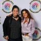 Two women stand side by side in front of a white backdrop featuring the circular logo and text “DECOLONIZED BEATZ INDIGENOUS WORLD PRIDE”. On the left, one woman wears a black blazer over a polka-dot shirt. She smiles warmly, standing with relaxed posture and her arm gently around the woman beside her. On the right, another woman is wearing a light purple blouse tucked into white pants with a decorative belt. She has long, styled hair and wears large hoop earrings and a necklace. She smiles with a playful tilt of the head.