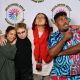Four individuals pose together in front of a white backdrop featuring a circular logo that reads “DECOLONIZED BEATZ INDIGENOUS WORLD PRIDE.” On the far left, a person with black hair tied up wears a green oversized shirt and light blue jeans. They’re smiling while resting their hand on Ritni Tears’ shoulder. Ritni, second from the left, is dressed in black with dramatic makeup and white contact lenses, striking a wide-eyed serious expression. Next is 9a, wearing a white t-shirt, gray shorts, and a red textured sweater draped around their neck. A camera hangs from 9a’s neck, and they’re smacking a kiss with their eyes closed, their arm wrapped around the fourth person. On the far right Mohammed Zahif is wearing a vibrant blue and red patterned outfit. He has short dark hair and a mustache, and is playfully posing with one hand holding a lollipop close to their face, looking to the side with their tongue slightly out.
