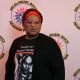 A person stands facing the camera in front of a white background with the DBIWP logo They wear a black t-shirt featuring a stylized portrait and bold red text that reads “Google me” Rayceen Pendarvis. Their accessories include red-rimmed glasses and a red and black headwrap. Their posture is upright and expressive, with a confident and charismatic presence