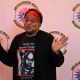 A person stands facing the camera with both hands raised, palms open and slightly angled outward in a gesture of emphasis in front of a white background with the DBIWP logo. They wear a black graphic t-shirt featuring a stylized portrait and the words “Google me” Rayceen Pendarvis in bold red lettering. Their accessories include red-framed glasses and a red head wrap. Their posture is upright and expressive, with a confident and charismatic presence.