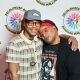 Two individuals stand side by side, posing closely for a photo, in front of a white background with the DBIWP logo. On the left, a person wears a patterned short-sleeve shirt, a gray cap with a white logo, and red-framed glasses. Their arm is wrapped around the shoulder of the person next to them, and they smile softly. On the right, a person wears a black headwrap with red patterns, red-framed glasses, a black cardigan, and a black graphic t-shirt with the text “Google me” Rayceen Pendarvis. Their head is rested at the other person’s shoulder.