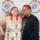 Two people pose in front of a white background with the DBIWP logo. Jen has short purple hair, they’re wearing clear glasses, bold red earrings and a strapless white dress with flower designs, her right hand is on their waist while the other embraces the other person, who’s wearing a black and red headscarf, round red glasses, a black t-shirt with a design and a black sweater. They both smile at the camera.
