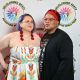 Two people pose in front of a white background with the DBIWP logo. Jen has short purple hair, they’re wearing clear glasses, bold red earrings and a strapless white dress with flower designs, her right hand is on their waist while the other embraces the other person, who’s wearing a black and red headscarf, round red glasses, a black t-shirt with a design and a black sweater. They both smile at the camera.