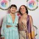 Two people stand side by side, smiling at the camera in front of a white background with the DBIWP logo. On the left, Tatiana is wearing a ruffled cutout baby blue dress with a V-neck, she has short hair and is wearing colorful beaded earrings, a necklace and white eyeliner, by her right side a woman with shoulder-length dark hair is wearing a long sleeved colorful patterned shirt, small feathery earrings, a pendant necklace and beige pants.
