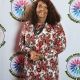 A woman poses in front of a white background with the DBIWP logo. She wears a long-sleeved, floor-length dress with a red, black, and white paisley pattern that flows elegantly to the ground. Her long, curly hair frames her face, and she smiles warmly at the camera. She accessorizes with a necklace and holds her hands gently together in front of her, standing with a poised and graceful posture.