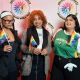 Three people pose in front of a white background with the DBIWP logo. Three individuals stand side by side, each holding a small rainbow pride flag. On the left, a person wears a black outfit with a long-sleeved top, black pants, a headband, and glasses. Their hair is braided and they stand with a relaxed posture, smiling softly. In the center, a person with short, curly hair wears a checkered coat over a black top, shirt and a beret. They hold their flag close to their chest and smile directly at the camera. On the right, a person wears a green and white shirt with “Pakistan” printed across the front, paired with dark pants and a plush toy clipped to their belt. Their hair is short and dark, and they stand with a casual, slightly angled posture, smiling with ease.