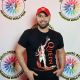 Tony stands facing the camera, in front of a white background with the BDIWP logo, wearing a red Adidas cap and a black T-shirt with a bold graphic of a giraffe and the word “QUEEN” in large red letters. He has a short beard. Their posture is upright and relaxed, with both arms resting naturally at his pockets and a soft, confident expression.