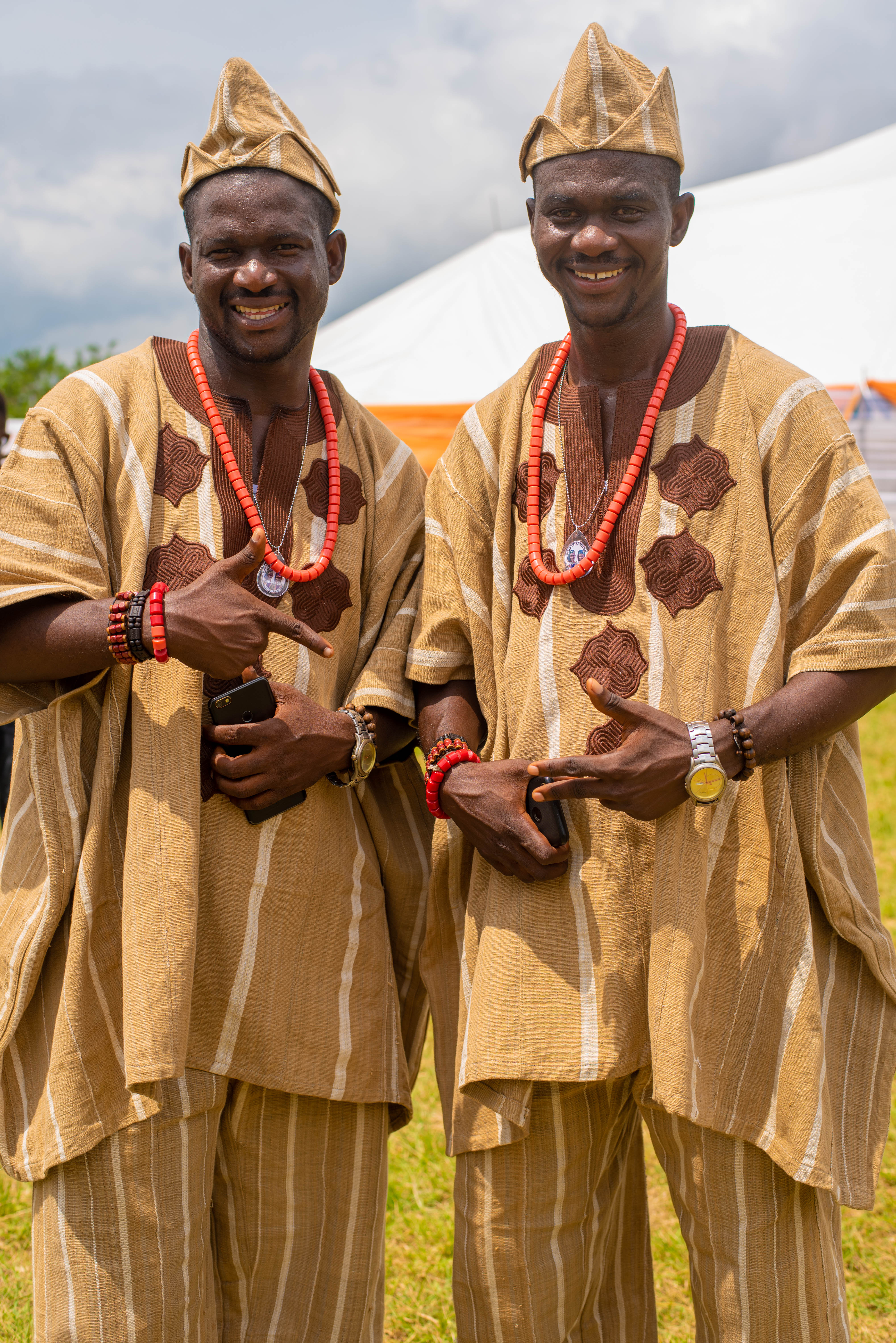 Taiwo and Kehinde Oguntoye, twin brothers and organizers of the World Twins Festival, wearing identical light brown agbada with striped patterns and red coral beads, standing outdoors during the World Twins Festival in Igboora, Nigeria. Their hands gesture playfully as they pose for the camera.