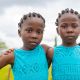 Two young twin girls standing close together with their arms around each other’s shoulders. They wear matching turquoise sleeveless dresses and gold earrings. Photographed during the World Twins Festival in Igboora, Oyo State, Nigeria.