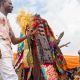 A masquerade figure dressed in a multicoloured patchwork robe and mask, holding ritual objects, is led by a man in white attire during a performance at the World Twins Festival in Igboora, Nigeria. Spectators and uniformed personnel can be seen in the background under tents.