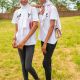 Two twin sisters stand side by side on grass, wearing identical white T-shirts, black jeans, white headscarves, and black slip-on shoes. Each carries a handbag, and one has her arm resting on the other's shoulder.