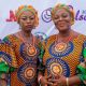 Two adult women wearing matching brightly colored Ankara dresses and headwraps stand in front of a promotional backdrop. Both have visible tribal facial markings and wear coral bead necklaces, with one smiling and the other maintaining a neutral expression.