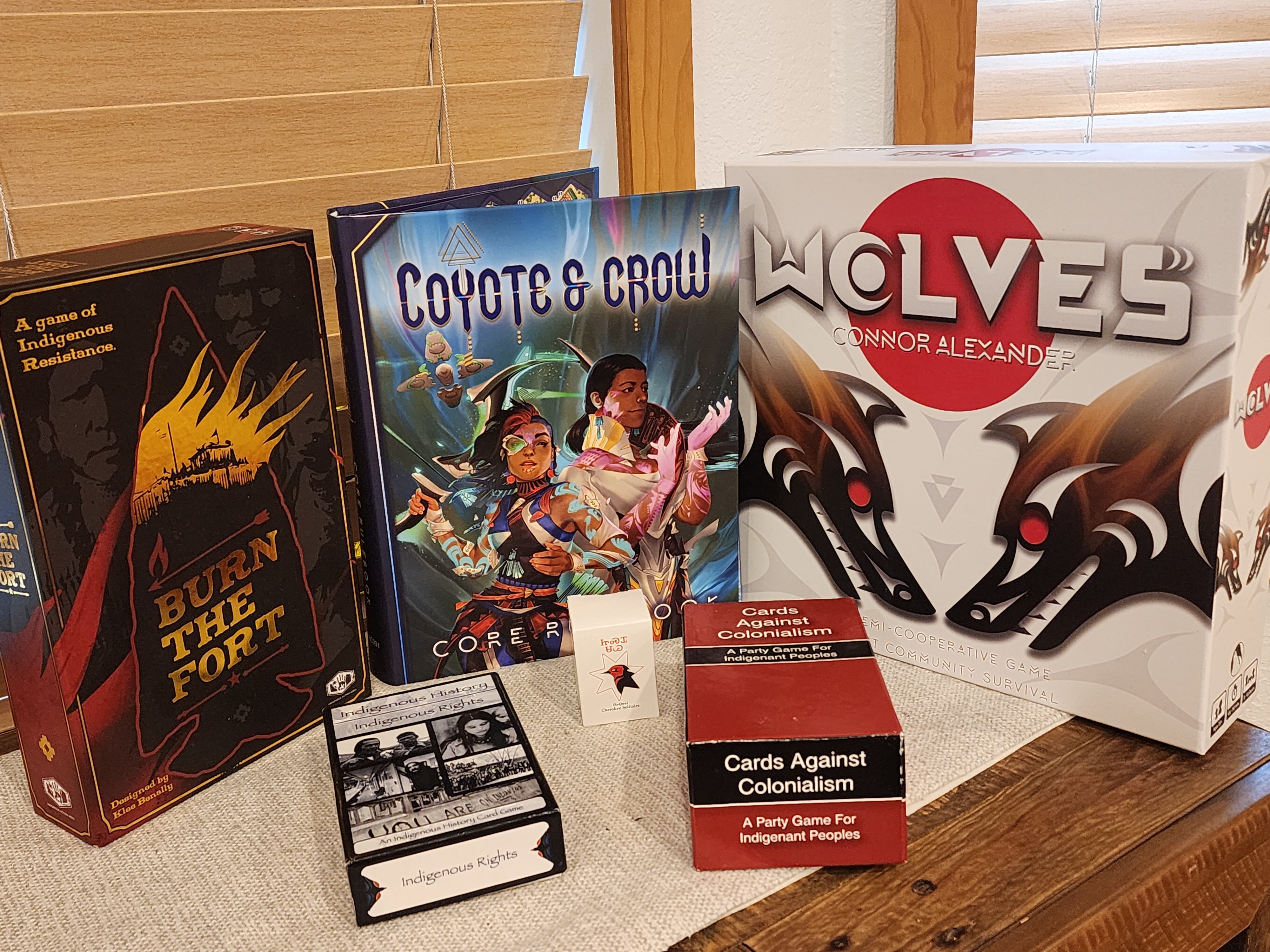 Board games are displayed on a white tablerunner on a medium dark wood surface, with a closed wooden blinds window in the background. From left to right: "Burn the Fort: A game of Indigenous Resistance," a dark-colored box with a burning fort illustration. "Coyote & Crow," a vibrant textbook featuring two Indigenous characters. "Wolves," a large white box with a red circle and two Coast Salish-style wolf heads. Three smaller card games: "Indigenous History, Indigenous Rights" and "Cards Against Colonialism: A Party Game For Indigenous Peoples” and “SaQuu (Cherokee Solitaire)”.