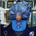 Chief Nike Davies-Okundaye stands inside her art gallery, the Nike Art Gallery in Lagos, Nigeria. She is dressed in rich indigo blue Adire attire, looking glamorous and hopeful. The background is softly faded, with canvas artworks hanging on the walls, highlighting the vibrant and creative atmosphere of the gallery she founded.