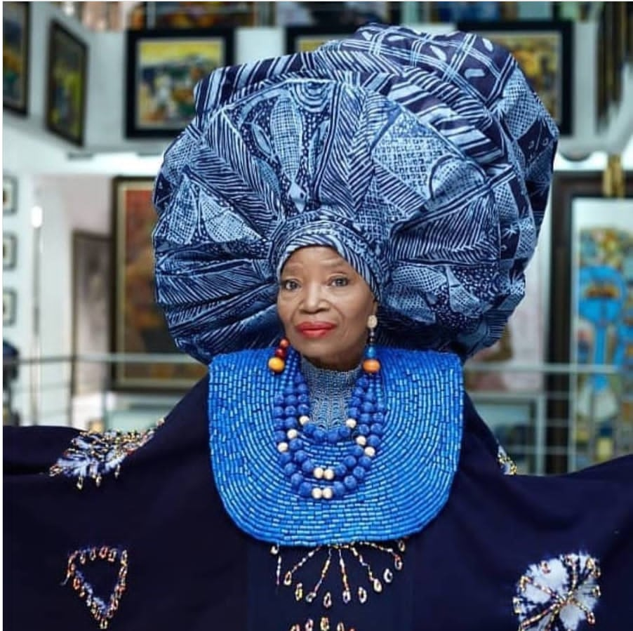 Chief Nike Davies-Okundaye stands inside her art gallery, the Nike Art Gallery in Lagos, Nigeria. She is dressed in rich indigo blue Adire attire, looking glamorous and hopeful. The background is softly faded, with canvas artworks hanging on the walls, highlighting the vibrant and creative atmosphere of the gallery she founded.