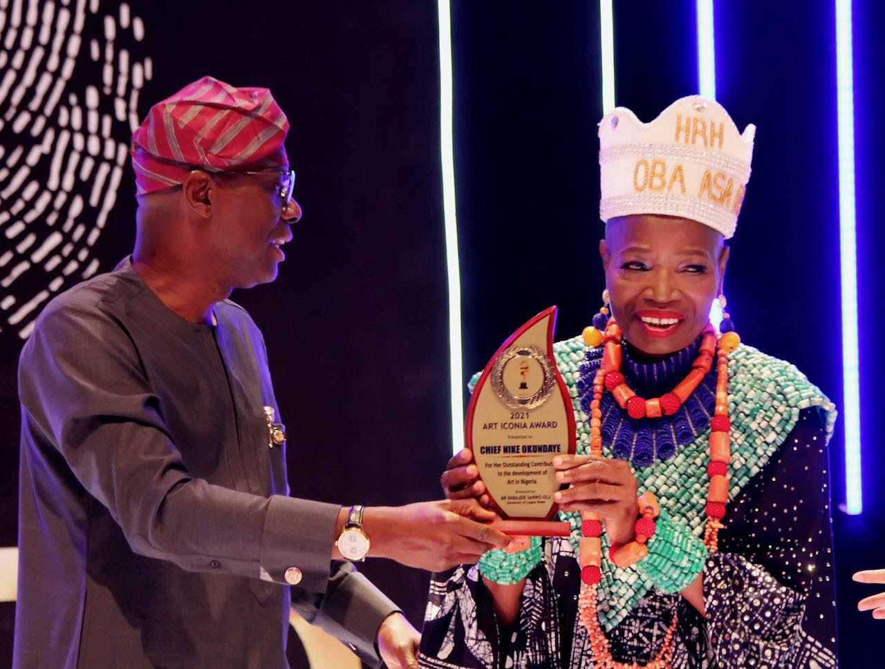 Chief Nike Davies-Okundaye stands at GAC Motors in Lagos, Nigeria, smiling warmly as she receives the Art Iconia Award from Governor Babajide Sanwo-Olu. Dressed in elegant shades of brown, red, and gold, she radiates happiness and pride. At 70, the renowned artist and cultural icon is honored for her lifetime dedication to promoting Nigerian culture through her vibrant artworks and artistic leadership. The moment captures both celebration and deep recognition of her enduring legacy.