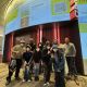A group of nine people wearing masks pose together in the lobby beneath a large curved digital projection advertising The Magazine anniversary event. The projection includes performer names, QR code, and event details. The group crouches and stands closely, smiling and making peace signs.