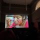 A large projection screen shows a close-up of an Indigenous Wayuu youth applying red paint to their cheek, with subtitles reading, “I am an Indigenous Wayuu person and I am bisexual.” Silhouetted audience members watch from darkened seating.