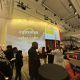 Guests gather in a spacious lobby with round black cocktail tables. A large yellow projection on the wall displays the Crushing Colonialism logo and event details. Some attendees eat, chat, or stand in groups