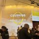 A close-up view of the Crushing Colonialism logo projected across a curved wall. People stand and socialize beneath it, some holding plates and cups. Lighting fixtures and ceiling rigging are visible above.