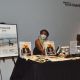 A person wearing glasses, a mask, and a black beret sits behind a table displaying The Magazine, La Revista, stickers, buttons, shirts, and informational flyers. A large framed poster of The Magazine stands on an easel to the left. A sign on the wall reads “Nancy K. Roche Chapel.”