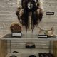 A glass display case contains Indigenous artwork. At the top hangs a large headdress made of fur, feathers, and antlers with a painted white mask. Below are a painted drum and an embroidered handbag in clear cases. The lower shelf displays Baltimore Center Stage merchandise including mugs, shirts, and a tote bag.