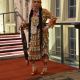 Denmi Whiteman stands on a red carpet wearing a traditional golden jingle dress decorated with silver cones and floral beadwork. They hold a black scarf with red flowers and wear long braids, a beaded headband, and colorful moccasins. The background shows tall windows and exposed white brick.