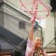 Rocka raises a cluster of interconnected pink hoops above her head, smiling broadly. She wears pink regalia with geometric beadwork. The image captures the motion and shape of the hoops.