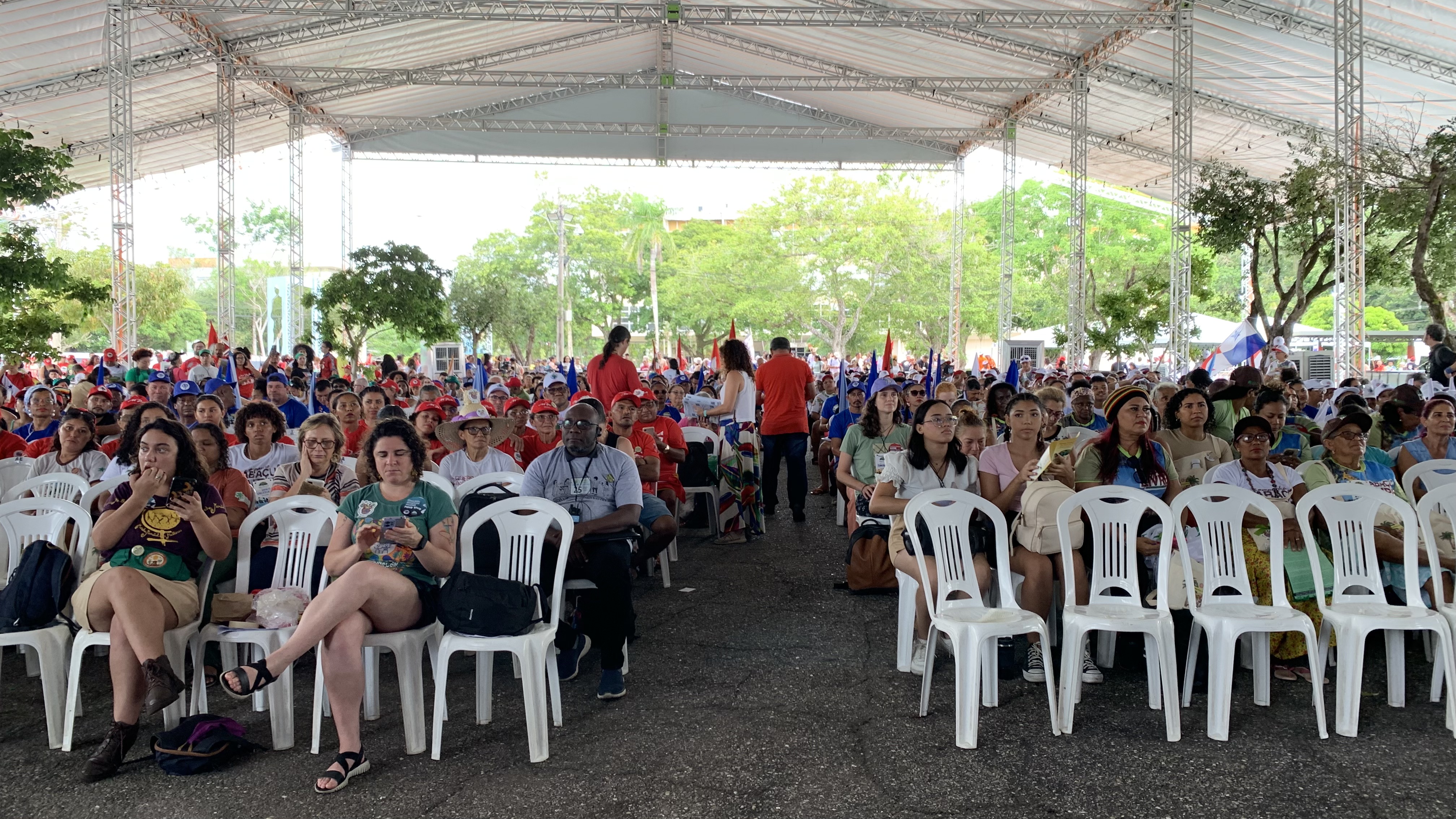 Dozens of participants sit on white plastic chairs inside a large pavilion at the Federal University of Pará in Brazil. Around them, colorful flags signal the diversity of movements present: blue flags represent fisherfolk and shellfish gatherers, red flags identify Brazil’s Landless Workers’ Movement (MST), and, to the right, banners mark the presence of Babaçu coconut breakers from Maranhão. Together, the raised flags reflect the broad range of social movements gathered in the space. Photo credit: Natalia Figueredo, November 2025.