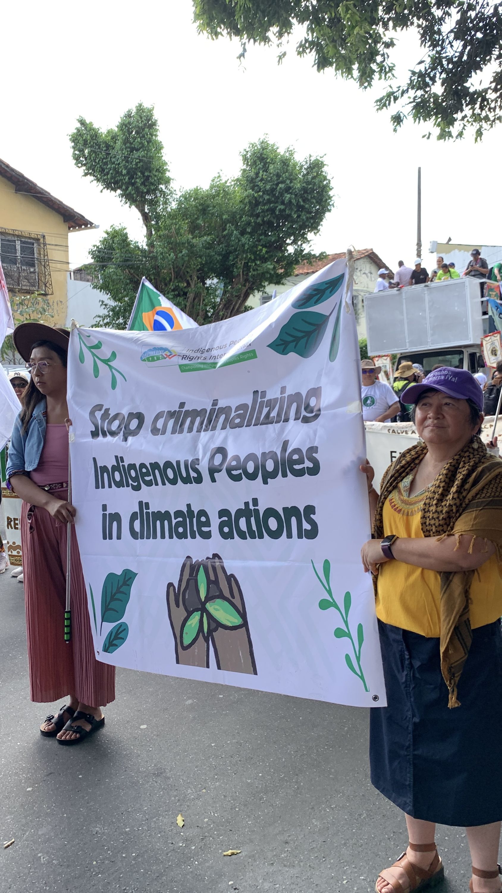 Two women stand side by side at a public demonstration, holding a white banner with green lettering that reads, “Stop criminalizing Indigenous peoples in climate actions.” One woman wears a pink skirt and a matching top, while the other wears a denim skirt and a yellow blouse. They face forward with a firm, determined stance amid the COP30 protests in Belém, Brazil.