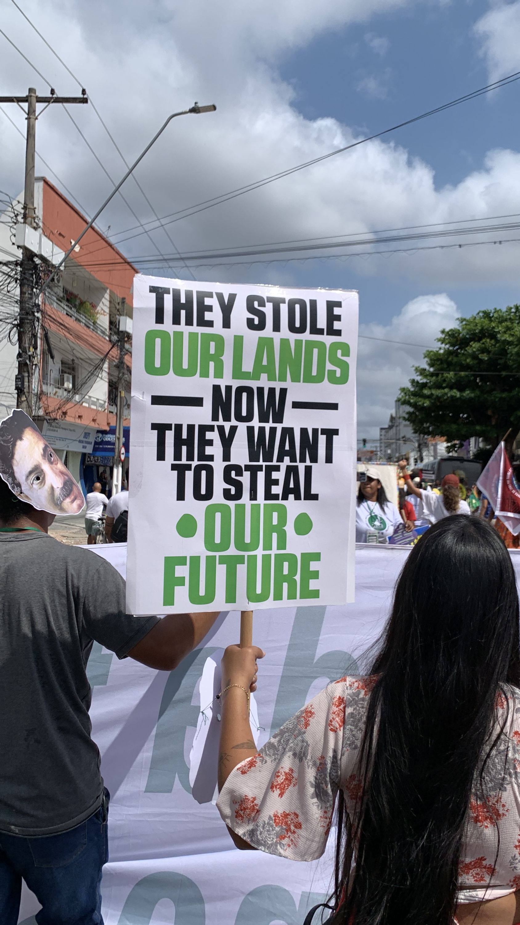 A woman holds a white protest sign with bold black and green lettering that reads, “They stole our land. Now they want to steal our future.” Beside her, another demonstrator wears a mask depicting Brazilian environmental activist and rubber tapper Chico Mendes, linking the protest to Brazil’s long history of land and environmental struggles.