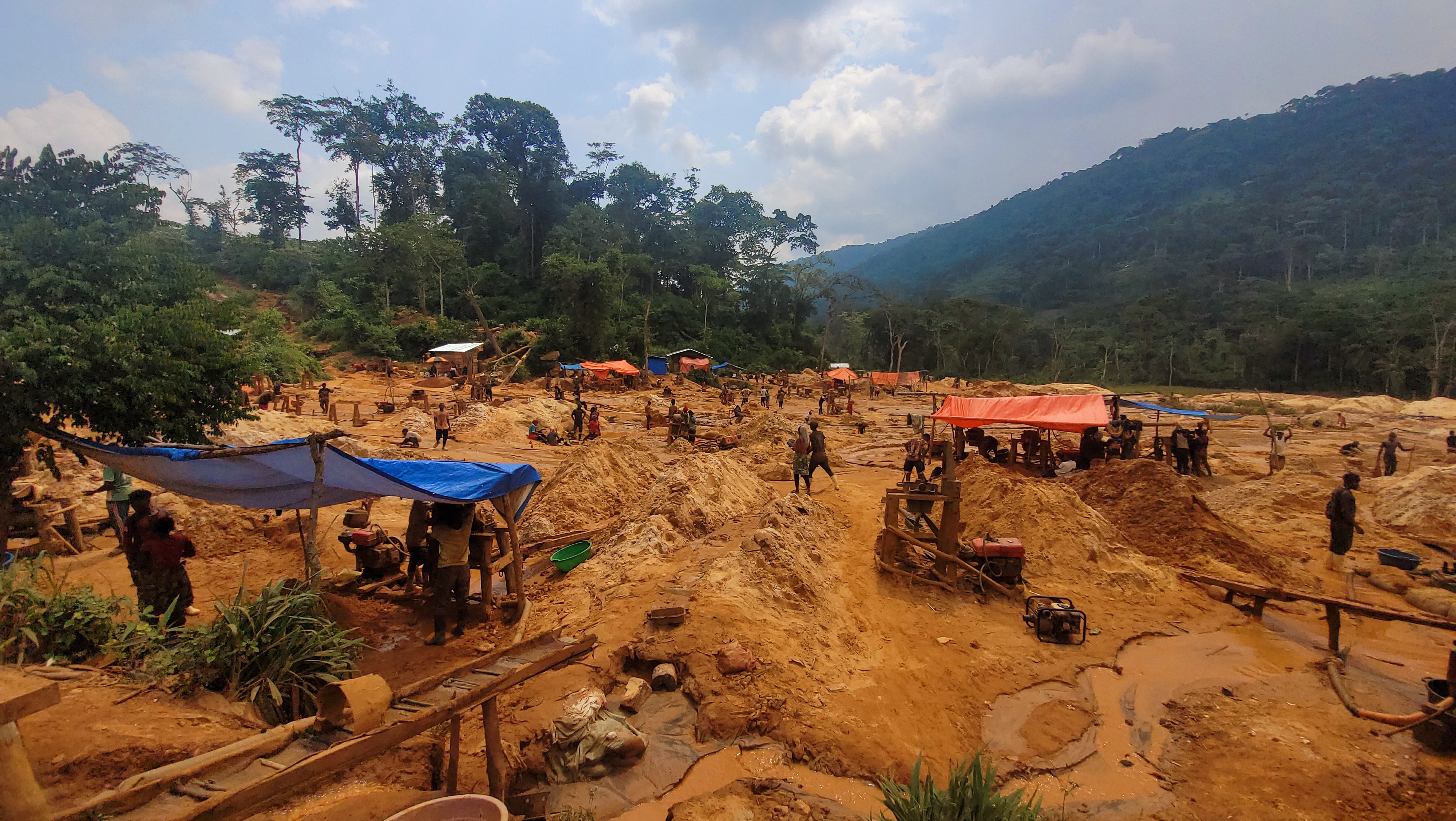 Wide view of the Pangoyi artisanal gold mining site in the Mangurujiba landscape, deep in the Congo Basin, where dozens of mostly young miners work across muddy, earth-toned pits carved into the forest floor. Makeshift wooden structures, blue and orange tarps, and small fuel-powered machines dot the site as miners dig, carry soil, and wash sediment, with dense green rainforest and surrounding hills rising in the background. Photo by Joseph Tsongo, July 30, 2025.