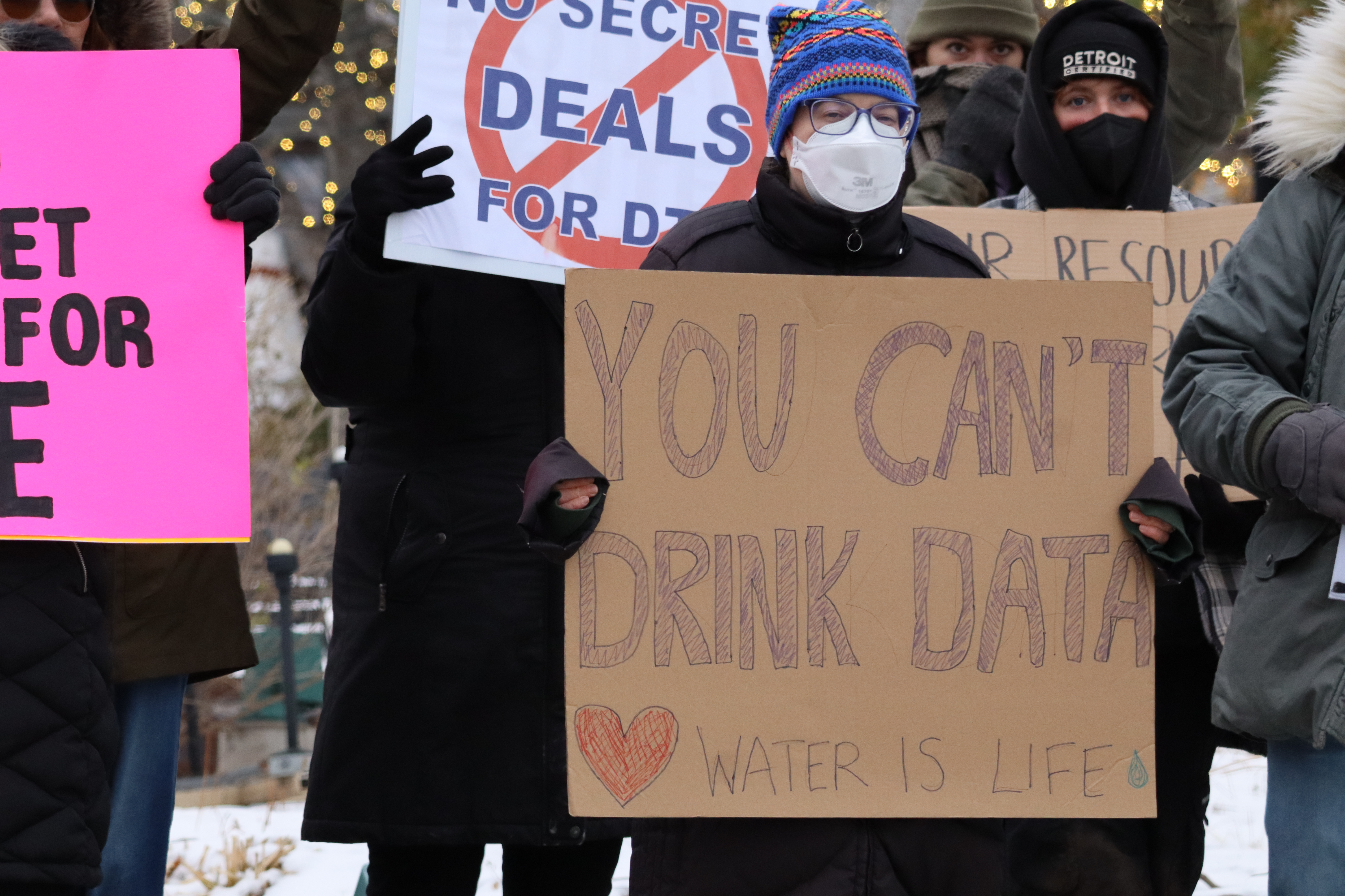 A group of bundled-up protesters stands outdoors in winter, wearing coats, hats, and face masks. In the center, a person holds a cardboard sign that reads “You Can’t Drink Data” with a red heart and the words “Water Is Life” written below. Behind them, others hold signs reading “No Secret Deals for Data.” Snow and holiday lights are visible in the background.