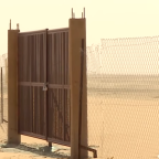 A sealed metal portal stands closed within a wire fence at a nuclear test site near Reggane. The gate is locked and reinforced with vertical metal bars, marking a fenced-off restricted area. The surrounding landscape is sandy, dry, and barren, with no people visible, emphasizing the site's isolation and controlled access. Photo credit: Tarek Hillali, January 2026.