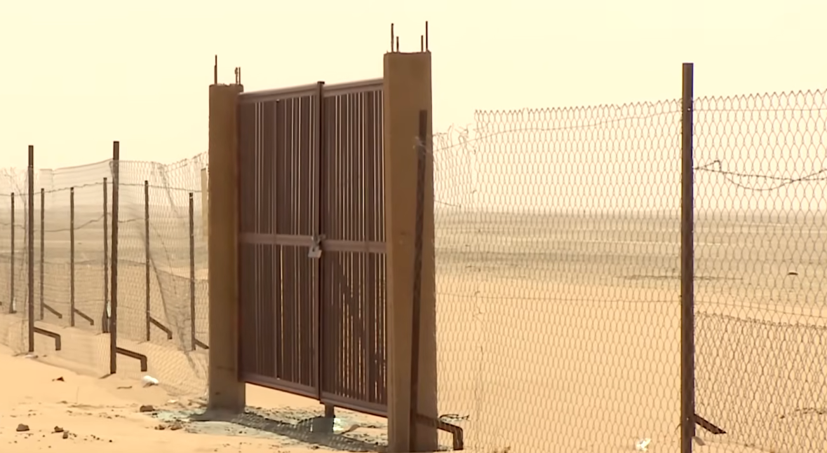 A sealed metal portal stands closed within a wire fence at a nuclear test site near Reggane. The gate is locked and reinforced with vertical metal bars, marking a fenced-off restricted area. The surrounding landscape is sandy, dry, and barren, with no people visible, emphasizing the site's isolation and controlled access. Photo credit: Tarek Hillali, January 2026.