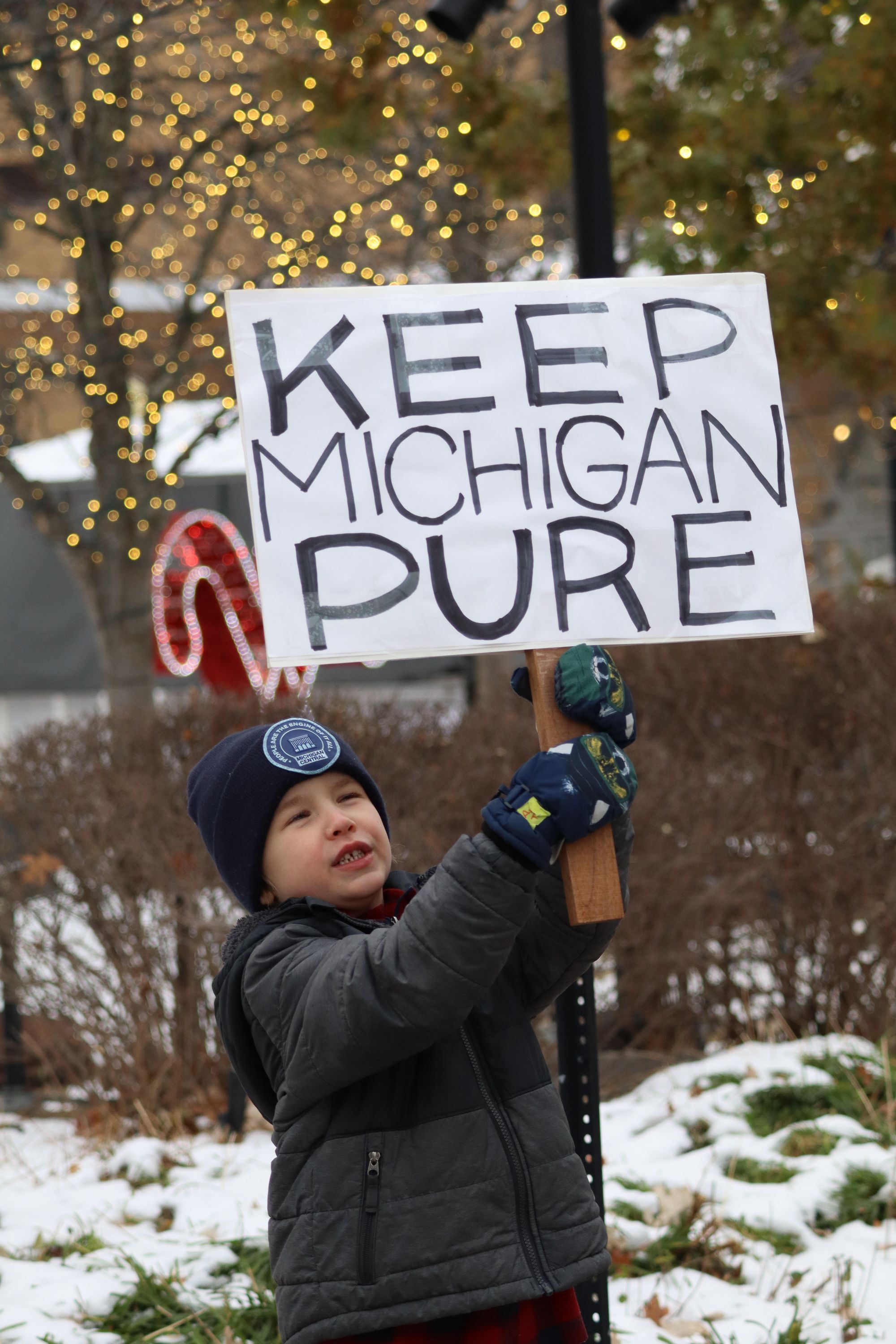 A child dressed in a winter coat, gloves, and a knit hat stands outdoors in the snow, holding a handmade sign above their head that reads “Keep Michigan Pure.” Holiday lights glow in the trees behind them, and patches of snow cover the ground.