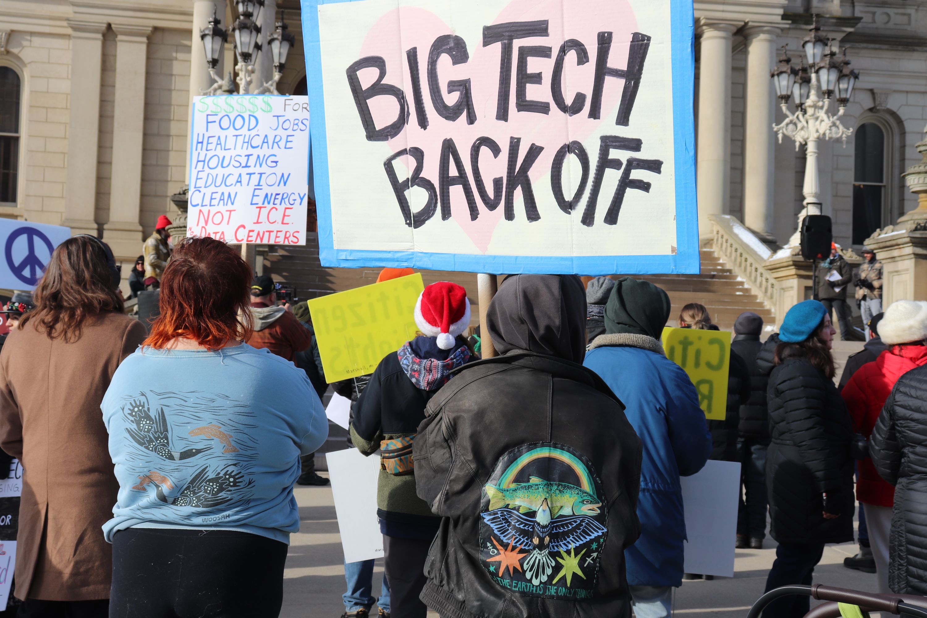 A group of protesters stands facing the steps of a government building. In the foreground, a large sign reads “Big Tech Back Off” with a pink heart in the background. Other signs in the crowd mention funding for food, jobs, healthcare, housing, education, and clean energy instead of data centers. People wear winter coats, hats, and scarves.
