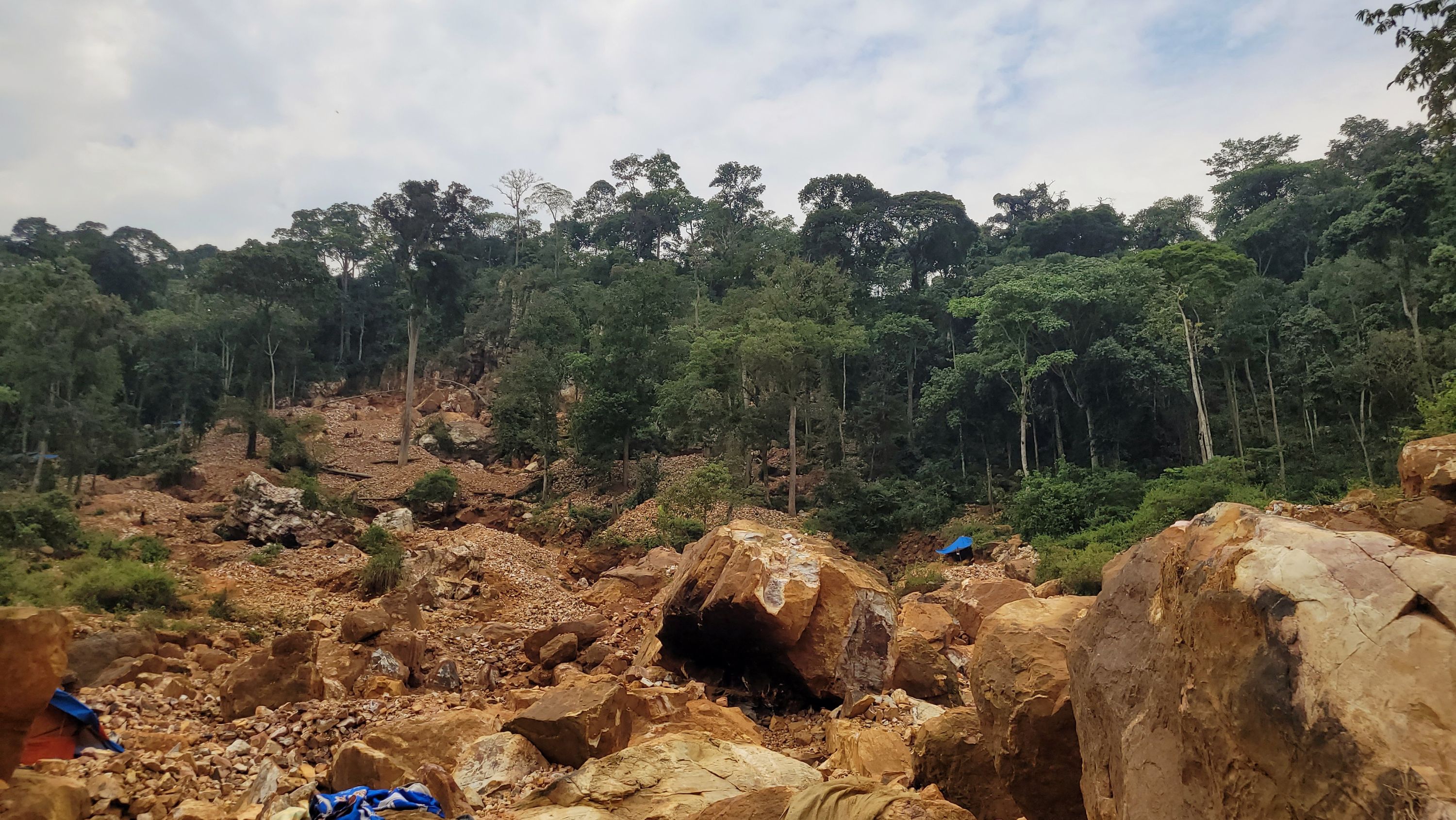 Rocky outcrops and scattered boulders dominate the Pangoyi artisanal mining area in the Mangurejiba landscape of the Congo Basin, where sections of forest have been cleared from a hillside. Exposed earth, broken stone, and mining debris fill the foreground, while patches of dense green forest remain standing in the background, highlighting the contrast between mined land and surrounding rainforest. Photo by Joseph Tsongo, July 30, 2025.
