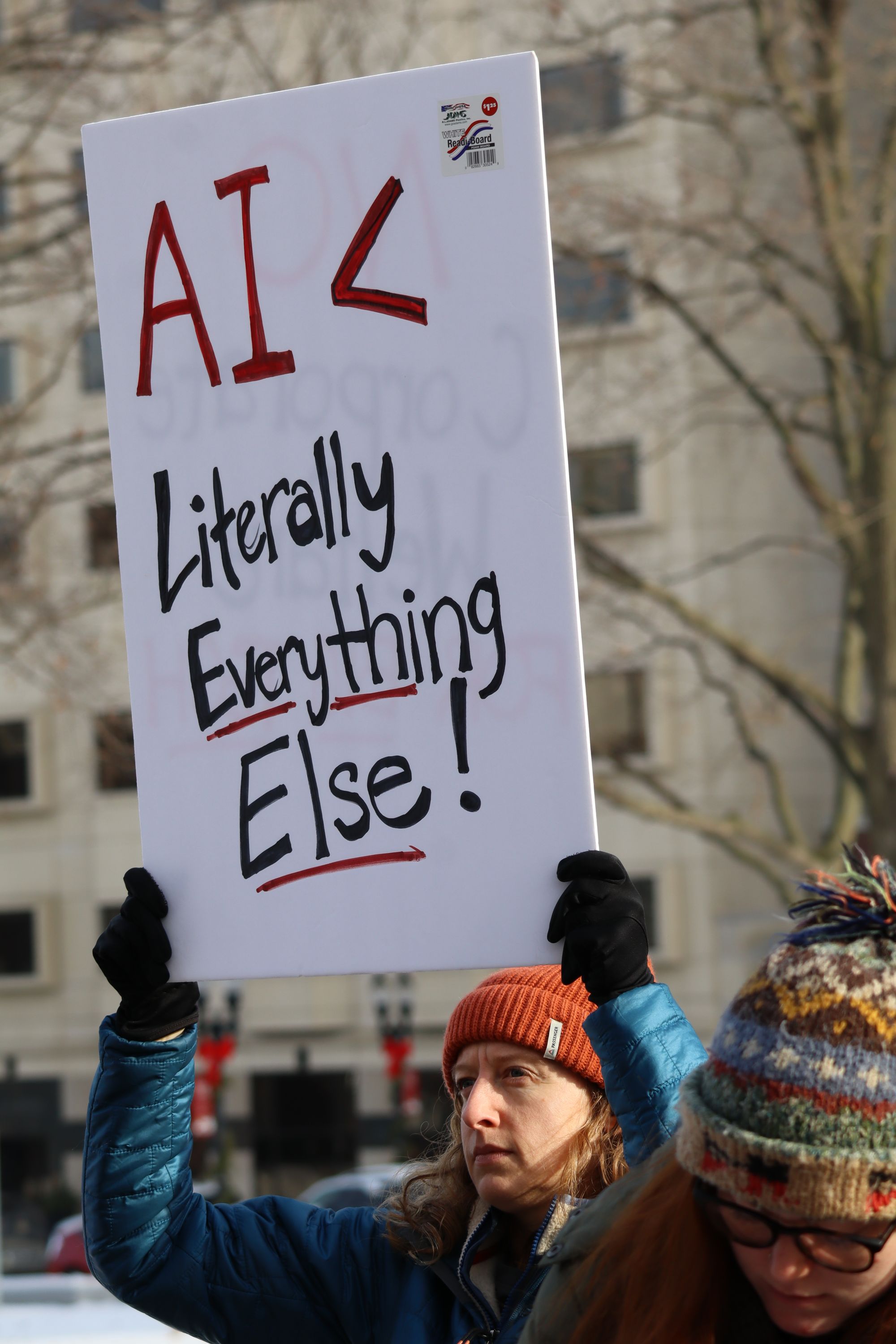 A person wearing a blue winter jacket, black gloves, and an orange knit hat holds a white sign above their head that reads “AI < Literally Everything Else!” in large red and black letters. Another person in a patterned winter hat stands beside them. A large building and bare winter trees are visible in the background.
