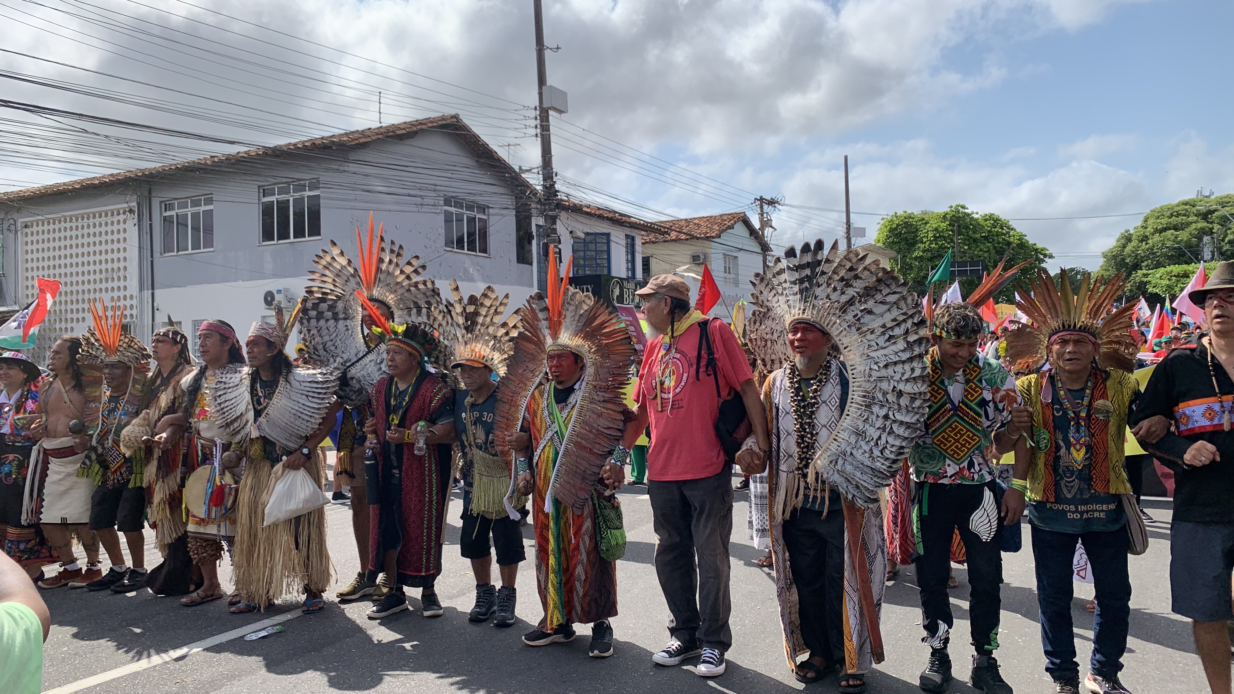 Approximately fifteen Indigenous representatives wearing traditional clothing and feather headdresses walk hand in hand down a city street during a march. They move forward together in a line, surrounded by other participants in the demonstration. Photo: Natalia Figueredo, November 2025.
