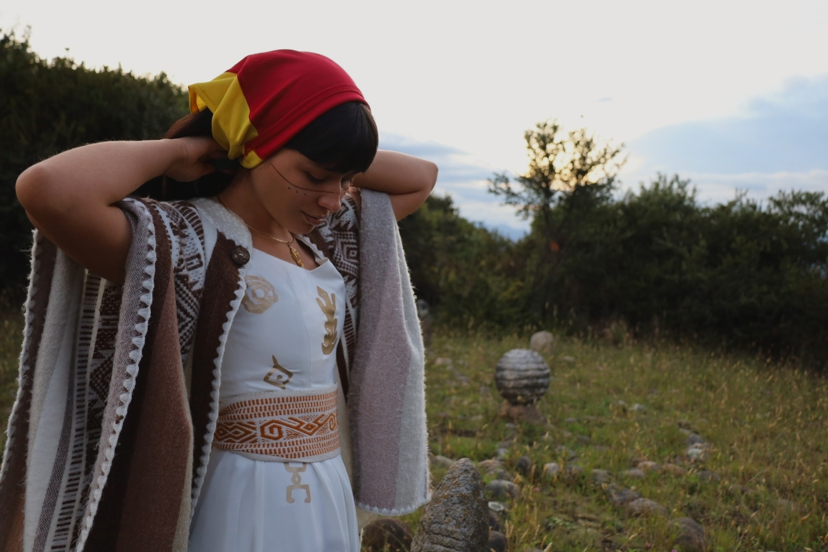 Lune stands outdoors at dusk, adjusting a red and yellow headscarf while wearing traditional clothing inspired by Muisca symbols, including a handwoven belt at her waist and a patterned wool mantle draped over her shoulders. Behind her, the open landscape and fading light frame a quiet moment of preparation and cultural reconnection.
