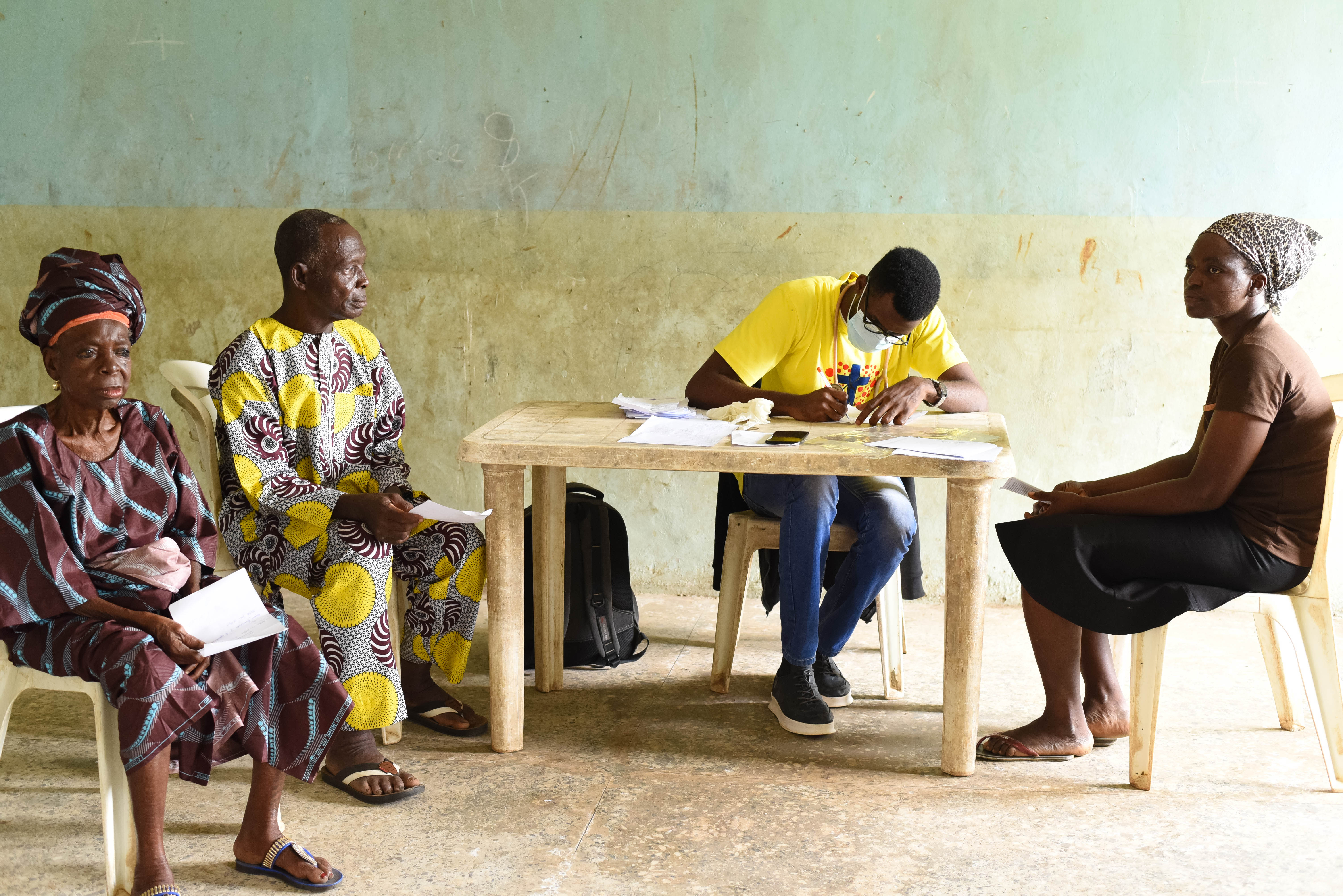 Inside a repurposed classroom in Langbin, an elderly resident, Mrs. Alice (left), and her neighbors wait for clinical consultations. The crowded waiting area reflects the high demand for care in a community where chronic health issues—ranging from waterborne infections to deteriorating vision—affect residents of all ages. Photo credit: Bukunmi Oyewole, December 11, 2020.