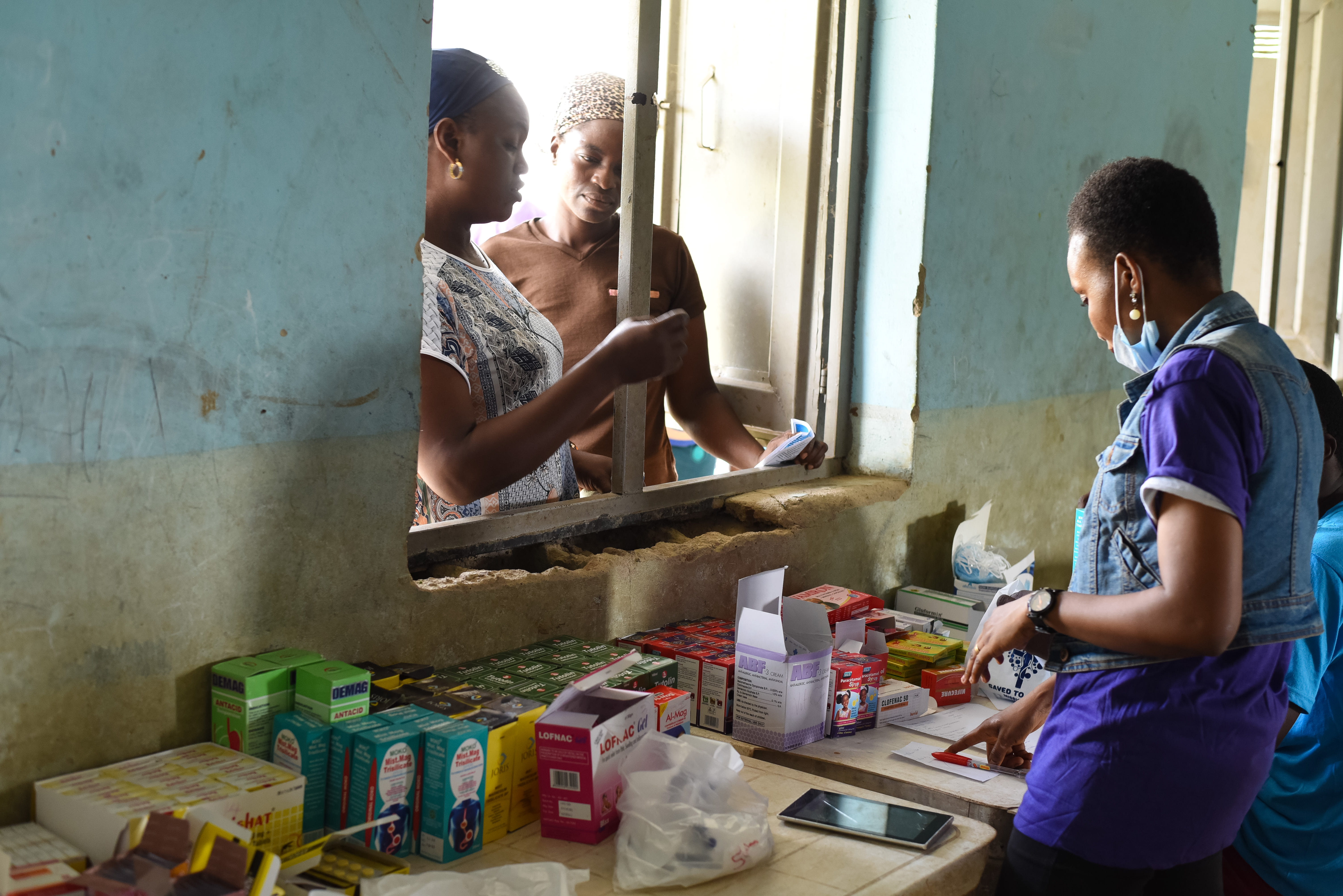 Mrs. Aderonke (left) and her neighbor wait at a makeshift pharmacy window to receive prescriptions and advice. This medical outreach in Ido Local Government serves as a vital bridge for families in Langbin who must otherwise manage chronic, water-borne health issues without local access to professional medical care. Photo credit: Bukunmi Oyewole, December 11, 2020.
