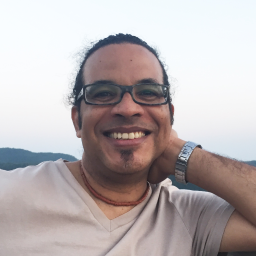Portrait of a smiling medium-brown-skinned man with his head resting on his right hand. He wears rectangular-shaped green and black acetate prescription glasses and sports a goatee with dark brown curly hair tied in the back. He has a Taino necklace made of small cylindrical clay beads and is wearing a beige V-neck t-shirt. Dusk sky and mountains are visible in the background.