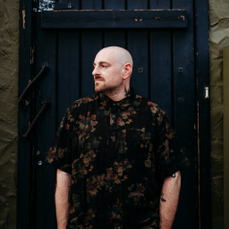 Light-skinned man with a shaved head and tattoos, wearing a dark floral shirt, standing pensively against a textured, dark wooden door, evoking a contemplative mood.